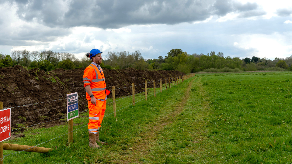 A construction worker wearing an orange high-visibility vest and blue hard hat stands near a fenced-off area in a grassy field under a cloudy sky. Signs indicate the area is closed.