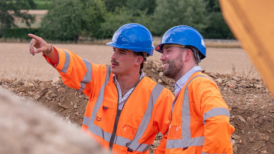 Two construction workers wearing blue hard hats and orange safety jackets, standing on a construction site. One worker is pointing towards a direction while the other is smiling and looking towards him.