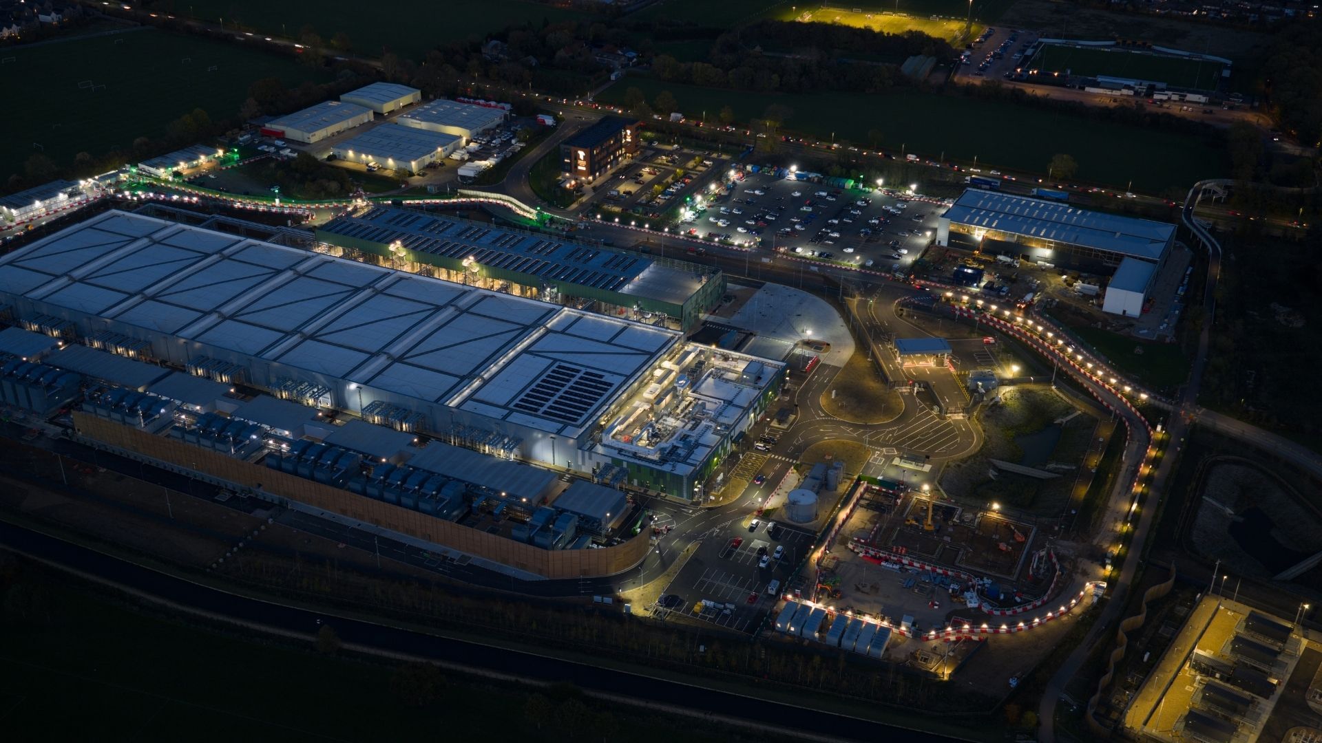 An aerial view of a large commercial complex at night, featuring a spacious building with a bright roof and illuminated parking lots. Surrounding areas are landscaped and lit, with various vehicles parked and pathways visible.