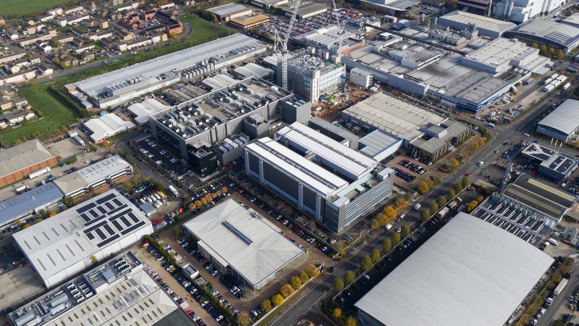 An aerial view of an industrial area featuring several large buildings and warehouses. The scene includes rooftops with solar panels, parking lots filled with vehicles, and a crane near a construction site, surrounded by greenery and residential areas.