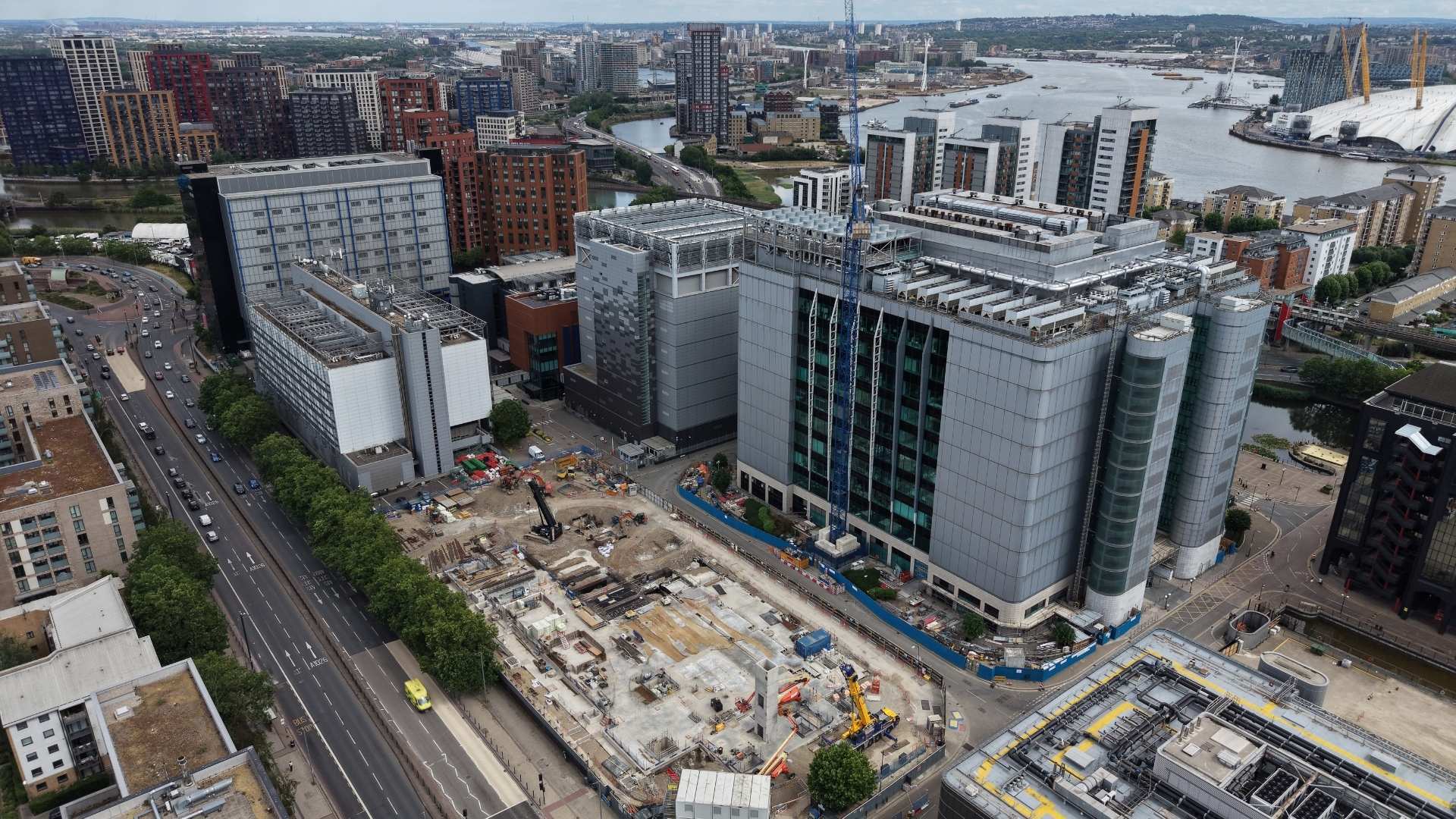 An aerial view of a construction site with several buildings, one featuring a glass facade, alongside a major road. The background shows urban developments and a river, with cranes and workers visible at the site.
