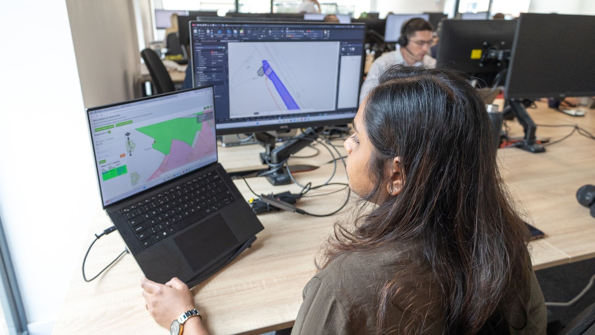 A woman with long dark hair sits at a desk, working on a laptop displaying a computer simulation. She is focused on the screen, which shows a colorful graphical representation. In the background, another person is seated at a workstation, working on their computer.