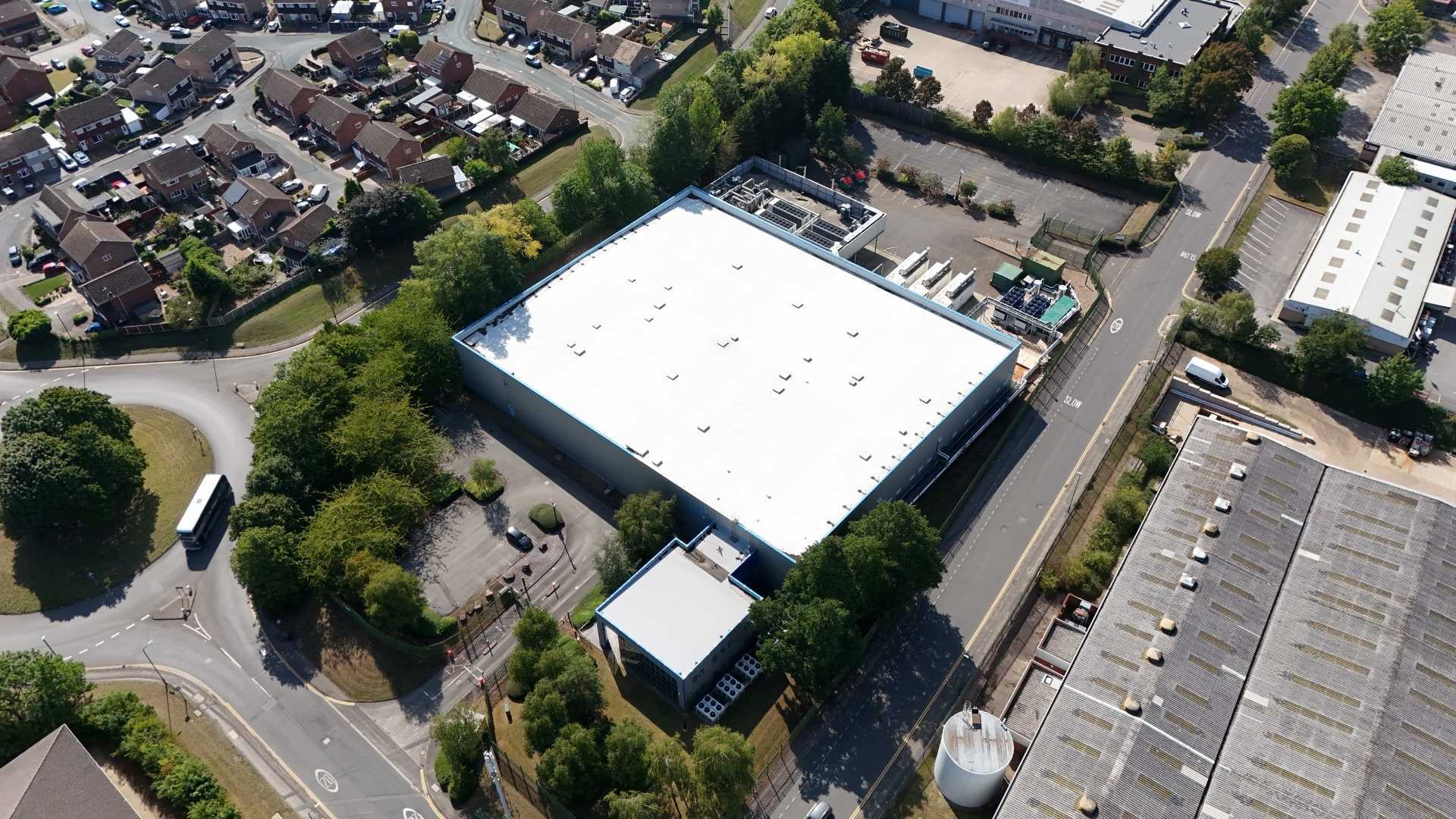 An aerial view of a large, flat-roofed commercial building surrounded by trees and a parking area. Nearby, there are residential houses and other industrial buildings visible along the road.