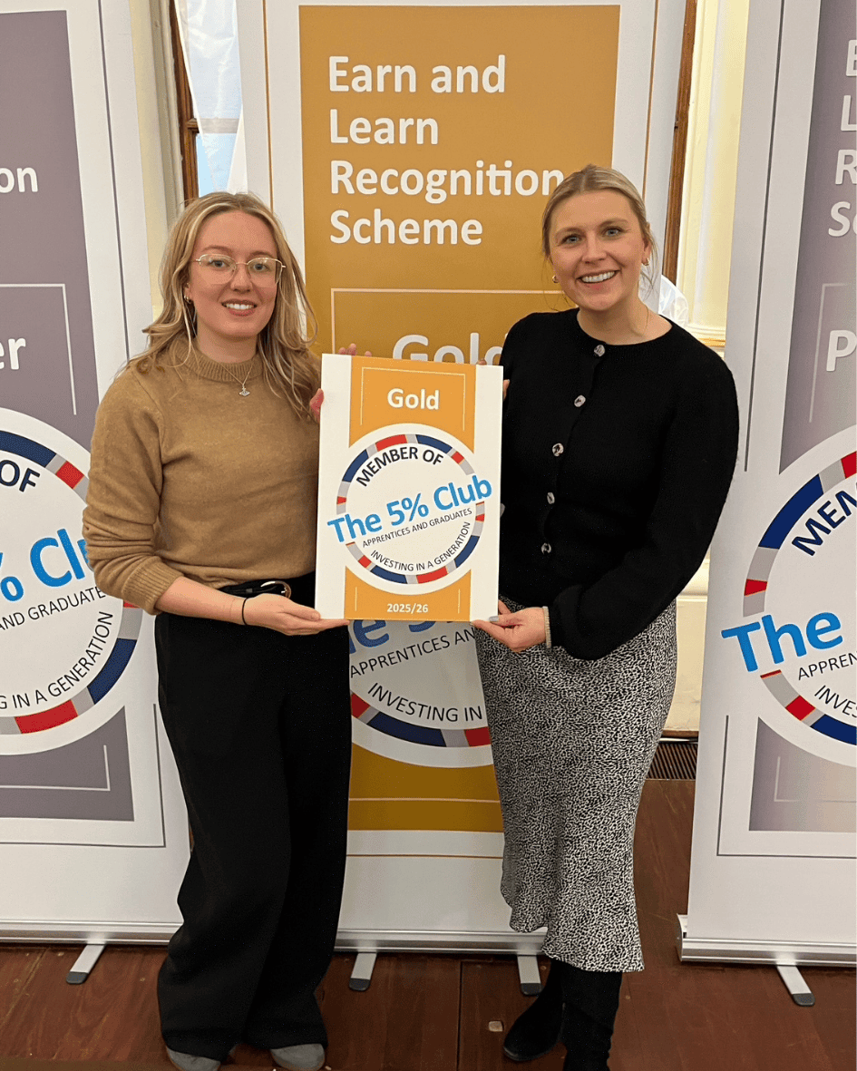 Two women standing together, smiling while holding a certificate titled 'Gold Member of The 5% Club' in a professional setting, surrounded by banners related to recognition and training.