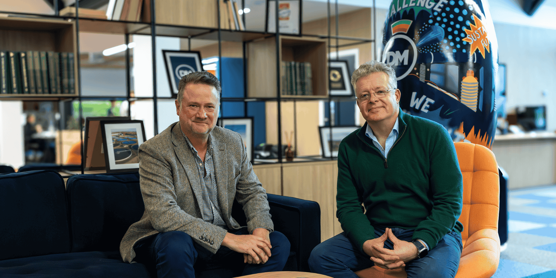 Two men sitting on a blue and orange couch in a modern office space. Behind them are bookshelves and artwork. One man in a grey blazer, the other in a green sweater, both looking at the camera.