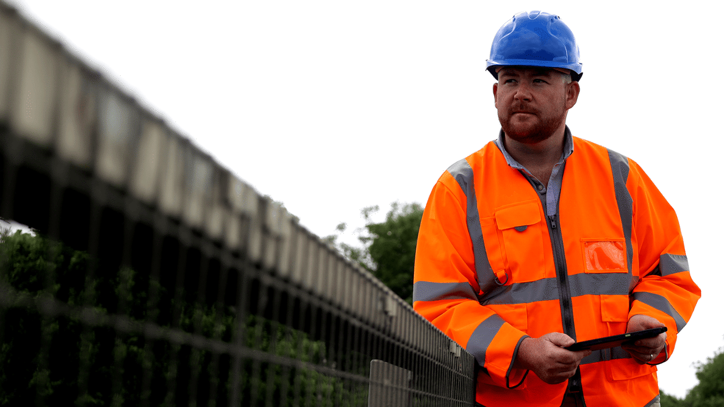 A man wearing a bright orange safety jacket and blue hard hat stands near a fence, holding a tablet. The background features greenery under a cloudy sky.