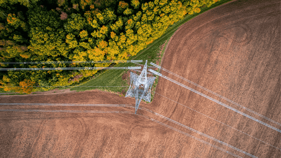 An aerial view of a power transmission tower located at the intersection of a dense forest and a cultivated field, showcasing power lines extending across the landscape.