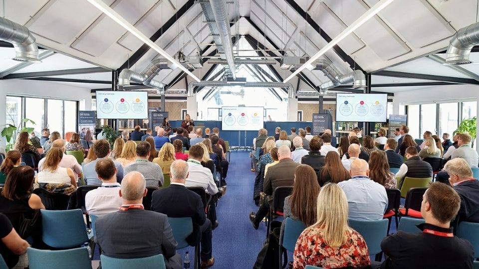 An audience seated in rows listens to a presentation in a bright conference hall with exposed ceiling beams and large screens at the front.