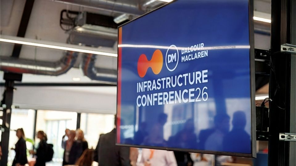 A digital display shows the 'Infrastructure Conference 26' logo for Dame Deborah Maclaren, with blurred attendees and ceiling pipes in the background.