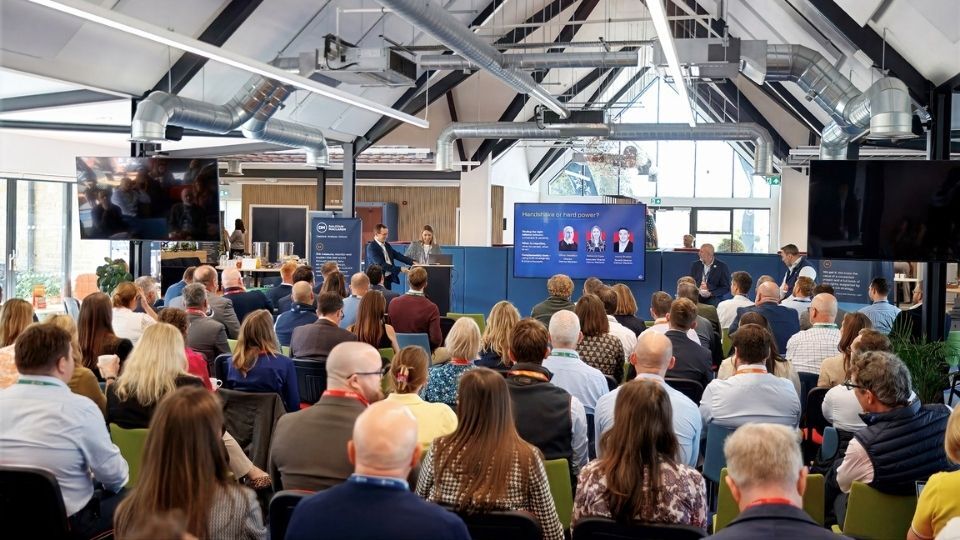 An audience seated in rows listens to a panel discussion on a stage, with large screens and exposed industrial beams in a bright conference hall.