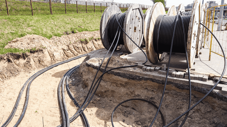 Two large wooden spools holding black cables located next to a trench in the ground. Several cables are uncoiled and laid out on the dirt surface, with green grass visible in the background.