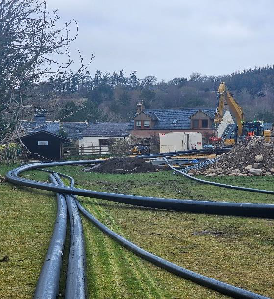 A construction site with large coiled black pipes running across a grassy area, surrounded by machinery and buildings in the background. Trees are visible in the distance under a cloudy sky.